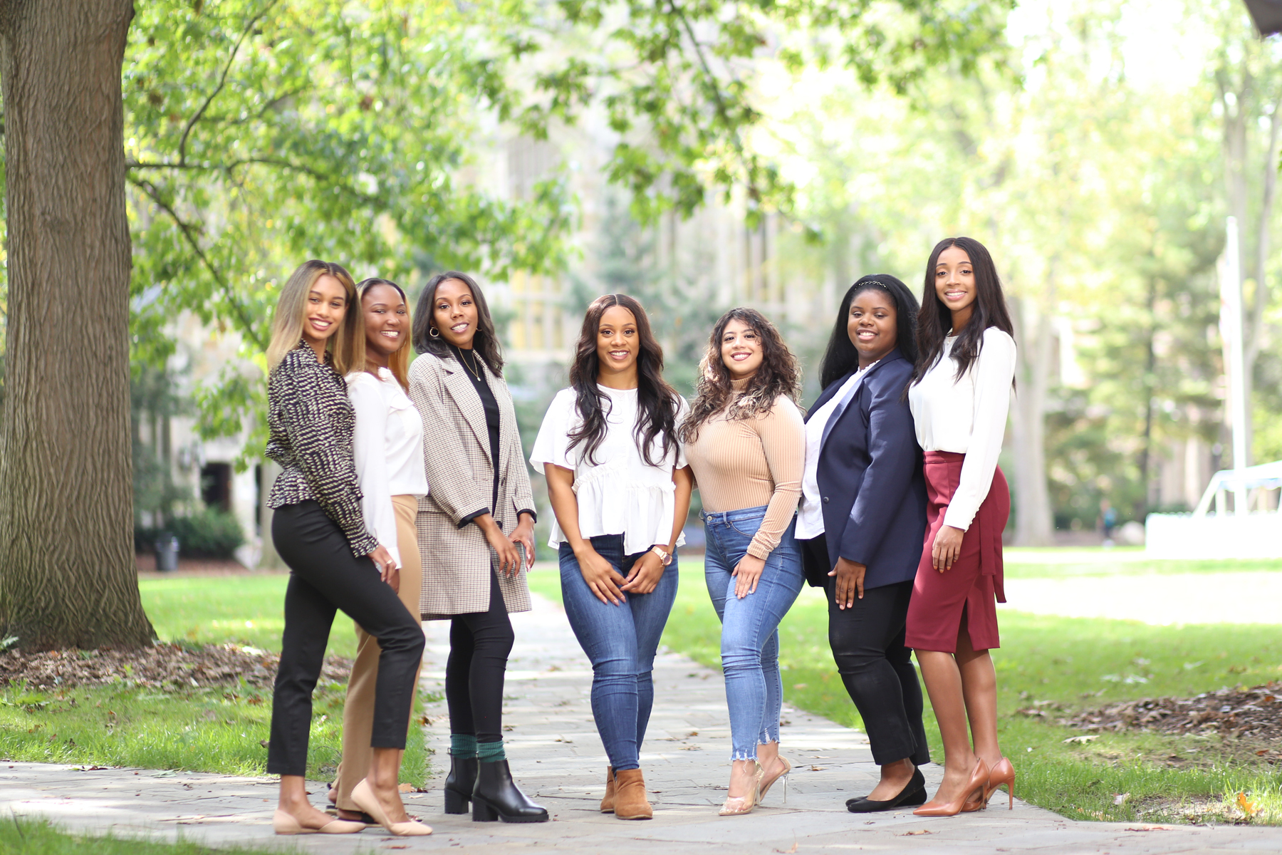 Seven women are posing together outdoors on a pathway, with trees and grass visible in the background.