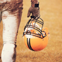 A football player in muddy gear holds a yellow helmet with a black stripe on a grassy field.