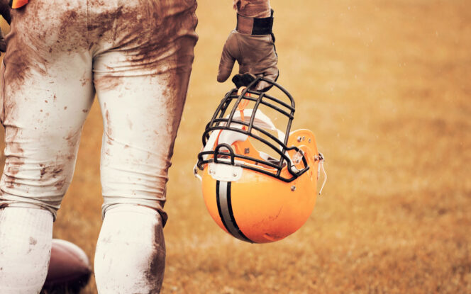 A football player in muddy gear holds a yellow helmet with a black stripe on a grassy field.