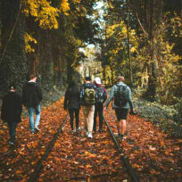 A group of people walks along a leaf-covered railroad track surrounded by autumn trees.