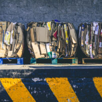 Three large bundles of compressed cardboard boxes sit on blue pallets against a gray wall, with a yellow-and-black striped curb in the foreground.