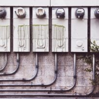 Six utility meters are mounted on a wall with green paint drips. Pipes run under the meters, and some greenery is present on the right side.