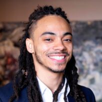 Smiling person with long, braided hair and a goatee, wearing a suit jacket, standing indoors.