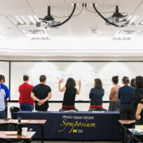 A group of people stand facing a large whiteboard covered in notes. A table with a cloth reading "Engaged Pedagogy Initiative Symposium" is in the foreground.