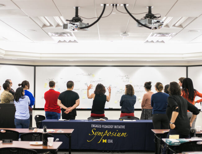 A group of people stand facing a large whiteboard covered in notes. A table with a cloth reading "Engaged Pedagogy Initiative Symposium" is in the foreground.