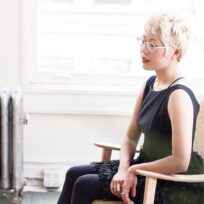 Woman with short blonde hair and glasses sits on a chair in a brightly lit room, wearing a black dress and dark leggings.