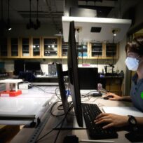 Person wearing a mask works at a desk with dual monitors in a dimly lit laboratory.