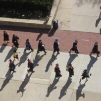 Aerial view of graduates in caps and gowns walking in rows across a paved area, casting long shadows.