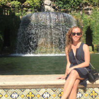 Woman in sunglasses sitting on a ledge in front of a fountain with cascading water and lush greenery.