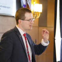 A man in a suit and tie speaks while gesturing with his right hand in a wood-paneled room with a wall-mounted screen and a standing lamp.