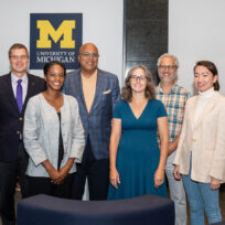 A group of eight people, five men and three women, standing indoors in front of a University of Michigan sign, smiling at the camera.
