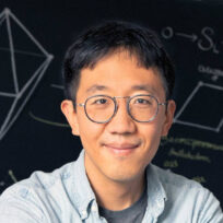 A person wearing glasses and a light blue shirt, standing in front of a blackboard with geometric drawings and equations.