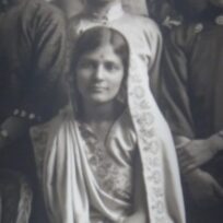 Black and white photo of a woman sitting, wearing a traditional sari with a bindi on her forehead, surrounded by others.
