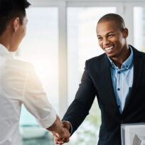 Two men shaking hands in a bright office setting, one holding a folder.