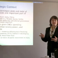 A woman in a suit stands near a presentation slide titled "Strategic Context," discussing governmental and public debt in various countries.