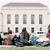 A group of people sit on a grassy area facing a large building with tall windows and a central entrance. Backpacks and bags are on the ground.