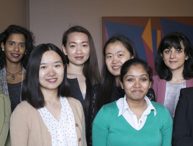 A group of eight women, some smiling, stands indoors in front of a painted wall.