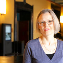 A woman with glasses and short hair stands indoors, wearing a blue top. She is in a well-lit hallway with soft yellow walls and dark fixtures in the background.