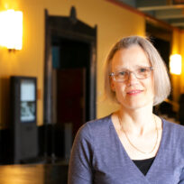 A woman with short hair and glasses stands indoors, wearing a purple top. The background features soft lighting and decorative elements.