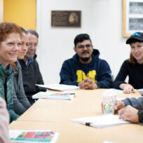 A group of people sit around a table in a meeting room, engaged in discussion. Papers and cups are on the table.