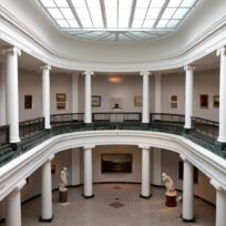 Spacious art gallery interior with white columns, a skylight, and various paintings and sculptures displayed on two levels.
