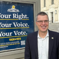 A man in a suit stands in front of a sign that reads "Your Right. Your Voice. Your Vote." The setting appears to be outside a building with steps and columns.