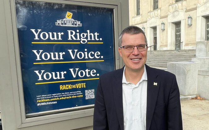 A man in a suit stands in front of a sign that reads "Your Right. Your Voice. Your Vote." The setting appears to be outside a building with steps and columns.
