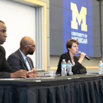 Three panelists seated at a table, one speaking into a microphone. University of Michigan logo displayed in the background.