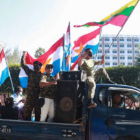 People in a pickup truck hold colorful flags, one wearing a mask, in a street with trees and buildings in the background.