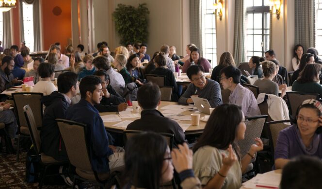 Large group of people seated at tables in a conference room, engaged in discussion and looking at documents and laptops under natural light from windows.
