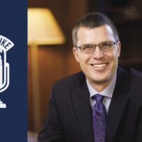 Man in a suit and glasses smiling, sitting indoors. Background shows a lamp and shelves. Logo with text "Open Mike" featuring a microphone is on the left.