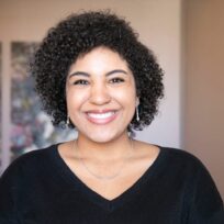 A person with curly hair smiles at the camera, wearing a black V-neck top. Two abstract paintings are on the wall behind them.