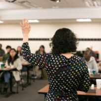 A person stands at the front of a classroom, raising their hand. Students are seated at desks facing the front, some with hands raised.