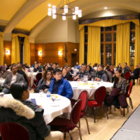 A man speaks on a stage to a large seated audience in a formal hall with yellow curtains and wooden walls. Tables are set with drinks and snacks.