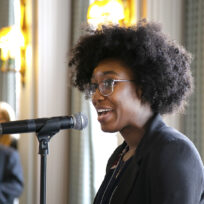 A person with curly hair and glasses speaking into a microphone in a room with people and lights in the background.