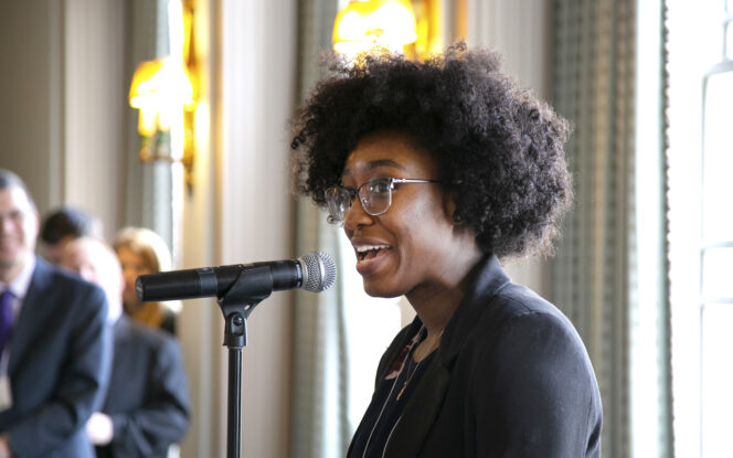 A person with curly hair and glasses speaking into a microphone in a room with people and lights in the background.