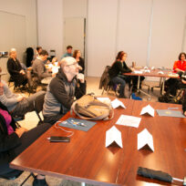 A group of people sit at tables in a classroom setting, attentively facing the front where a speaker or presentation is likely taking place.