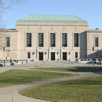 A large, rectangular, stone building with a green roof, flanked by barren trees, and surrounded by a grassy area and walkways under a clear blue sky.