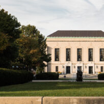 A large, rectangular building with columns and multiple windows surrounded by trees and greenery under a partly cloudy sky.
