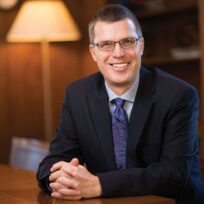 A man in glasses and a suit sits smiling at a wooden table in a room with a lamp and bookshelves.