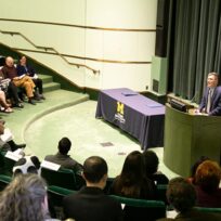 A man stands at a podium speaking to an audience seated in an auditorium. There are two tables covered with blue cloth on the stage.