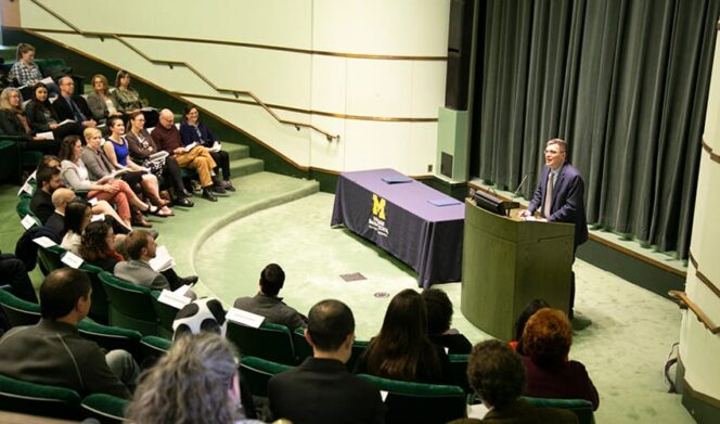 A man stands at a podium speaking to an audience seated in an auditorium. There are two tables covered with blue cloth on the stage.
