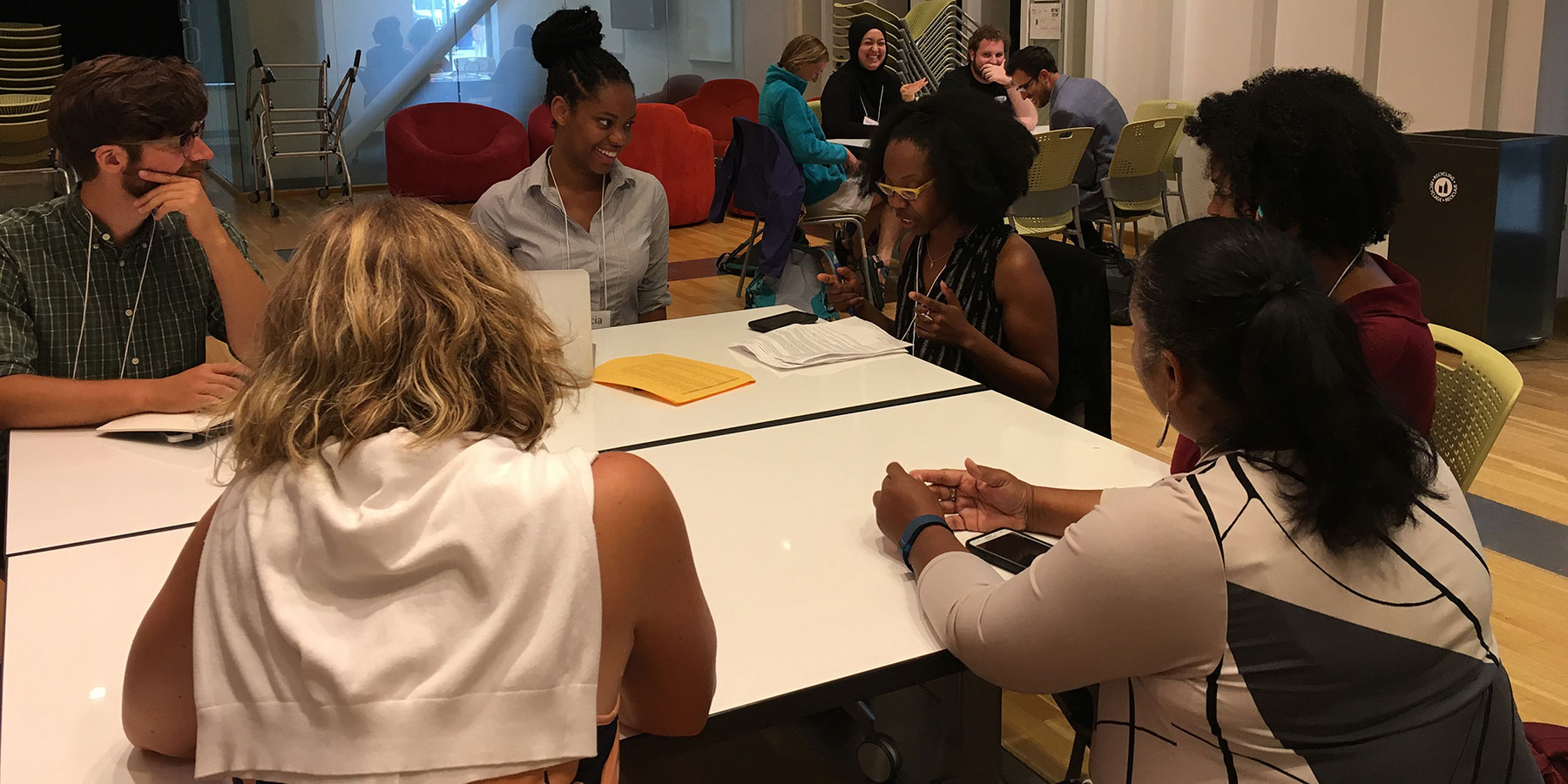 A group of six people sit around a table in a meeting room, three facing the camera, two with backs turned, and one in profile. Notes and documents are visible on the table.