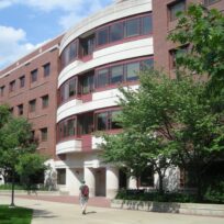 A person walks past a multi-story red brick and white stone building with large windows, surrounded by leafy trees, under a partly cloudy sky.