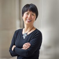 A person with short hair smiles while standing with arms crossed, wearing a black top and a necklace with circular pendants, in front of a neutral blurred background.