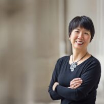 Person with short black hair, wearing a black top and necklace, smiling with arms crossed. Blurred background.