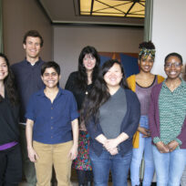 A diverse group of eight people standing indoors, smiling at the camera, with a simple office background.
