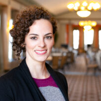 Smiling person with curly hair in a sweater and black cardigan stands in a large, elegantly lit room with tables and chairs.
