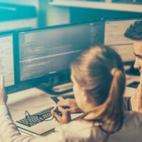 Two people collaborate at a workstation with multiple monitors displaying code and documents, while a calculator and a pair of glasses sit on the desk.