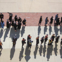 A group of graduates in caps and gowns walk in a procession on a concrete pathway, casting long shadows on the ground.